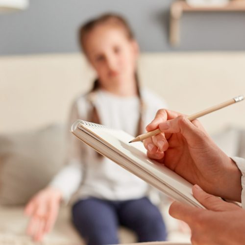 Female child having session with psychologist who takes notes on her emotional and mental state, carefully listening to the girl's concerns and issues.